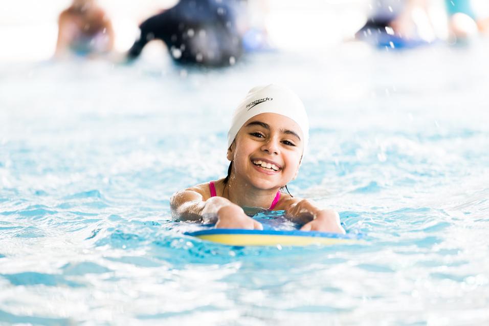 Lächelndes Mädchen mit weißer Badekappe schwimmt auf einem Schwimmbrett in einem Pool. Spritzendes Wasser ist sichtbar.